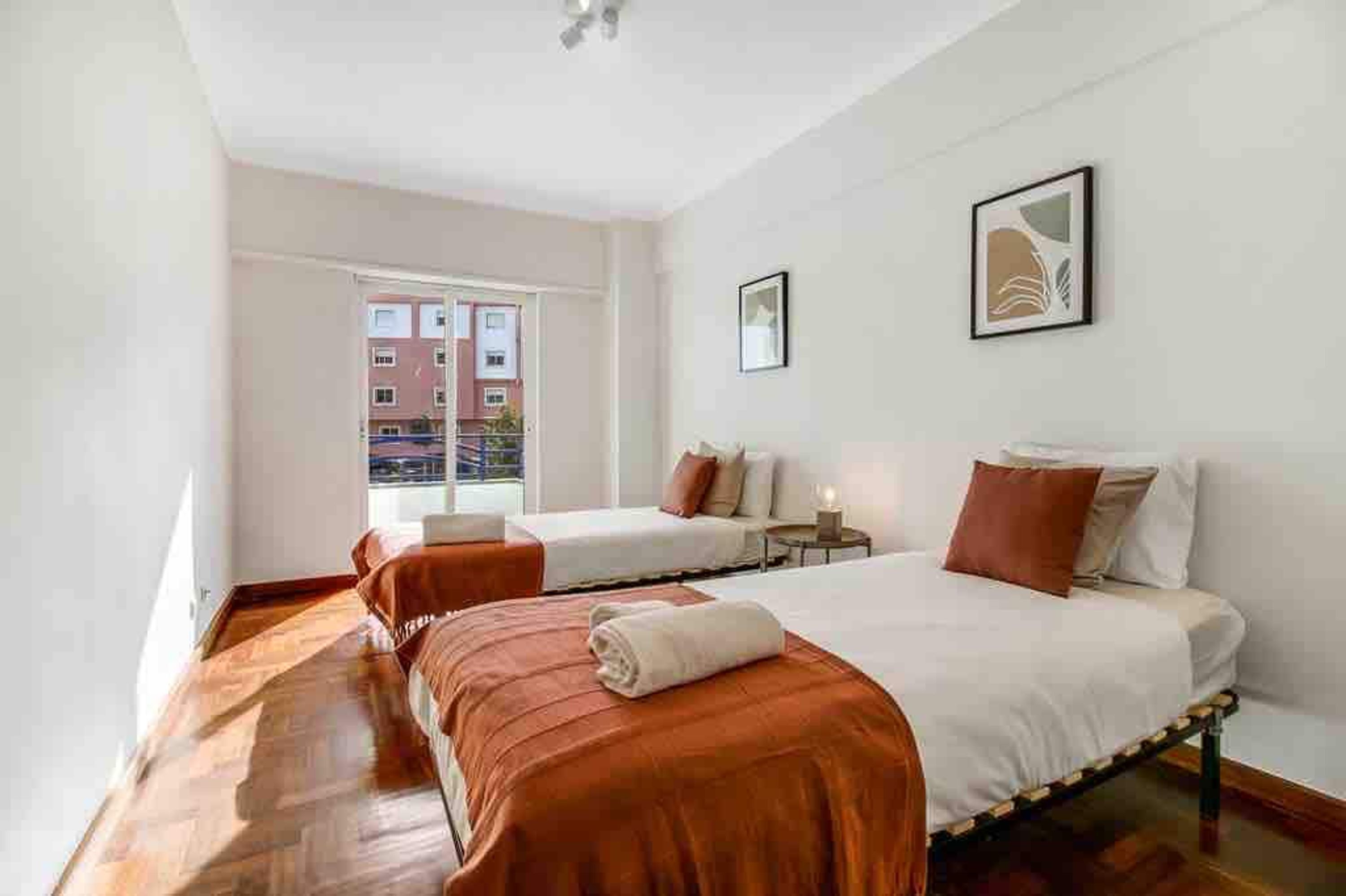 A hotel room with two beds, a small table, and a window with a view of buildings.