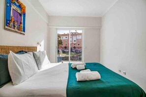A hotel room with a bed, pillows, a green bedspread, a window with a view of buildings, and a door.