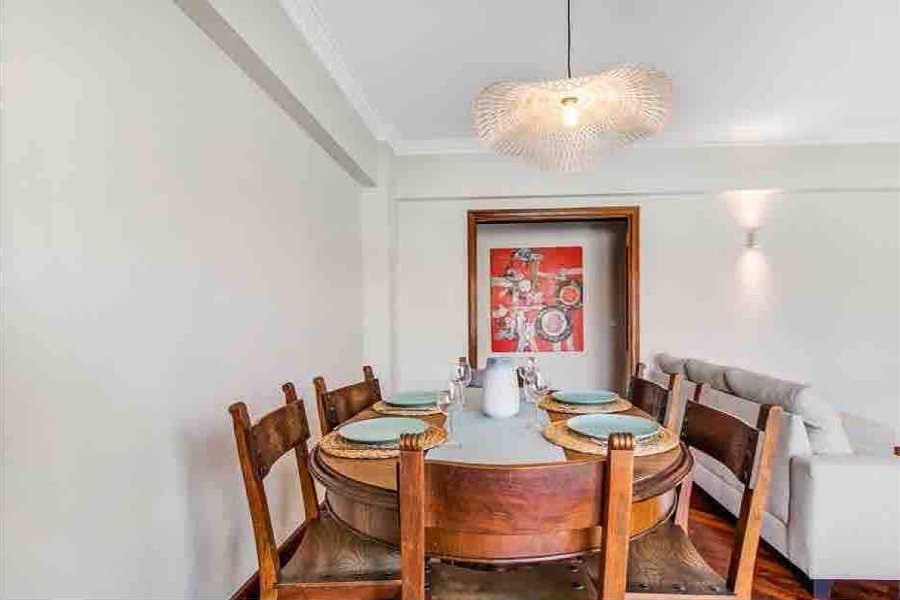 A dining area with a round table, wooden chairs, a hanging light fixture, and a framed artwork on the wall.
