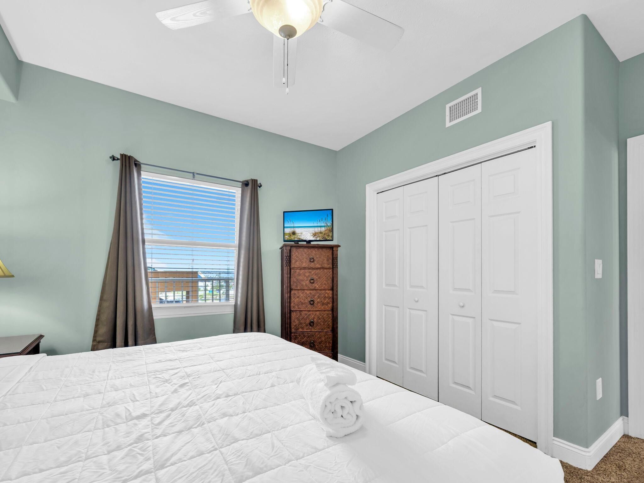A bedroom with a bed, a dresser, a ceiling fan, a window with curtains, and a closet.
