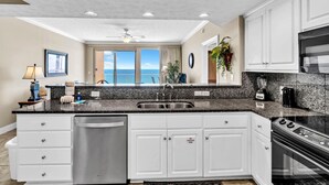 A modern kitchen with white cabinets, a granite countertop, a dishwasher, a stove, and a view of the ocean through large windows.