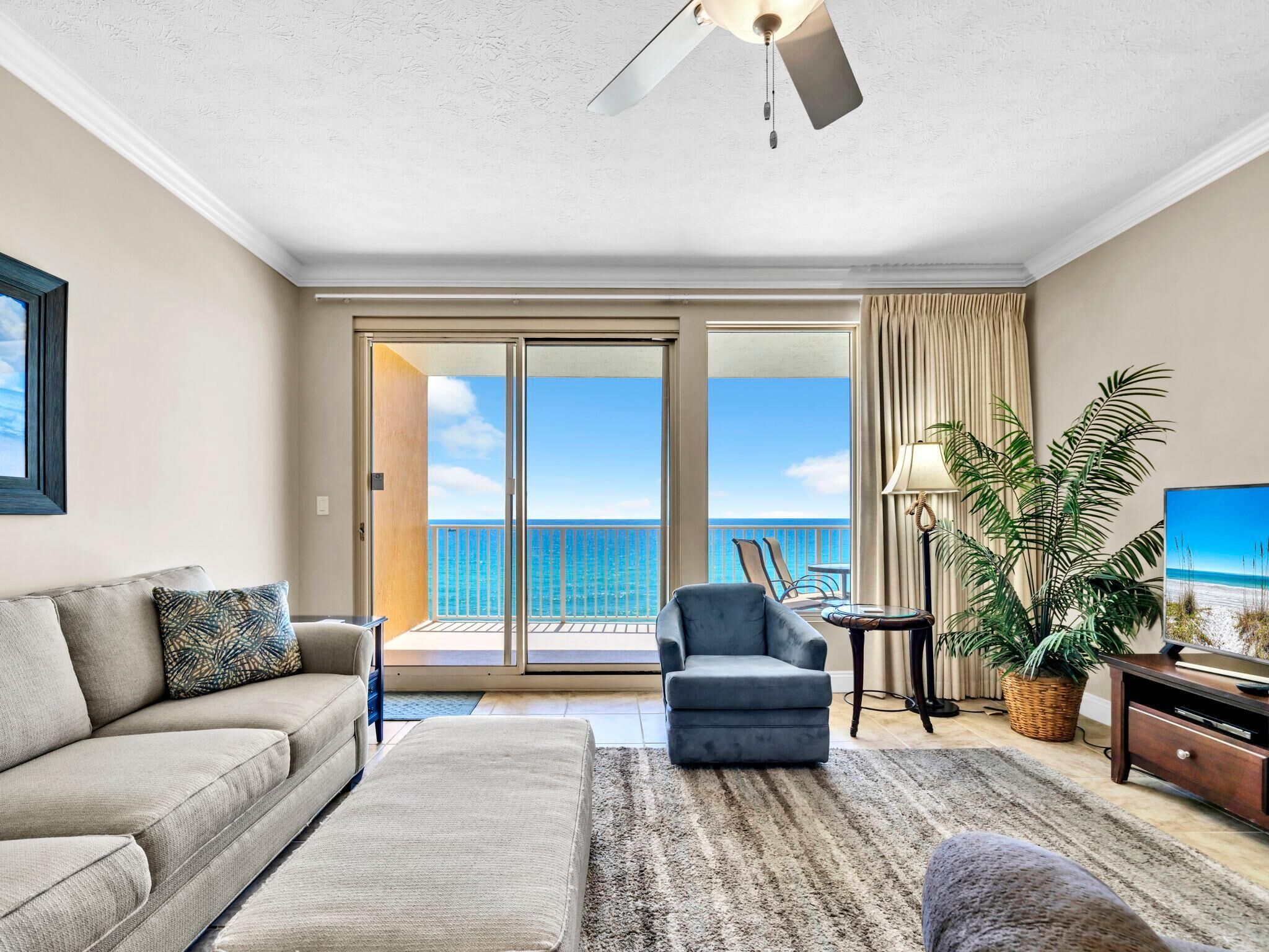 A spacious living room with a balcony, a ceiling fan, a sofa, a chair, a TV stand, and a potted plant.