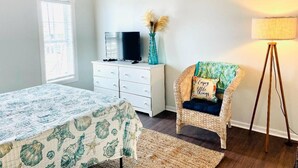 Quarto com cama, penteadeira, televisão, cadeira de vime e luminária de chão.