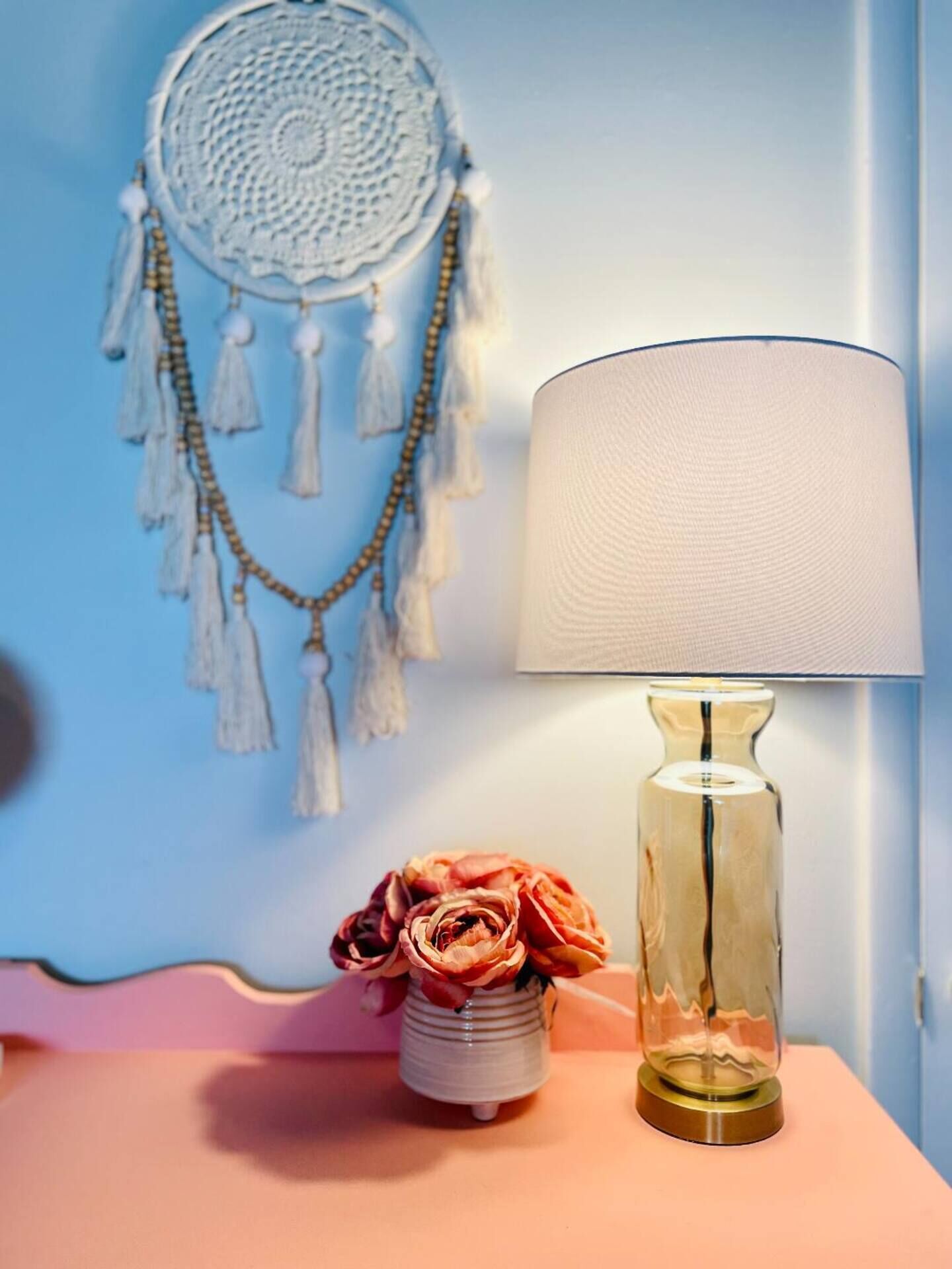 A bedside table with a lamp, a dreamcatcher, and a vase with flowers.
