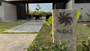A modern building with a stone sign reading "AUDAZ" in the foreground, a patio area with a table and chairs, and a view of a palm tree and sky. - Casa Audaz: Private Pool, Jacuzzi & Daily Cleaning in a Peaceful Escape (Circasia)