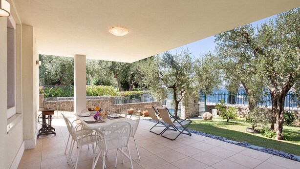 A covered patio with a table and chairs, a small table with a vase and flowers, and a view of the sea and trees. - Apartment Righini with Pool (Torri del Benaco)
