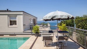 A poolside area with a view of the sea, a lounge chair, an umbrella, and a small table. - Apartment Righini with Pool (Torri del Benaco)