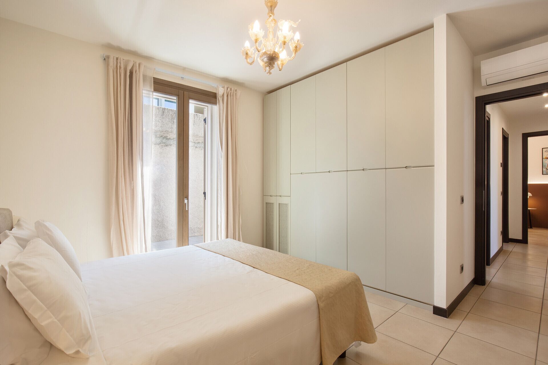 Une chambre à coucher avec un lit, une armoire, un lustre et une vue sur l’extérieur, visible à travers une porte vitrée.
