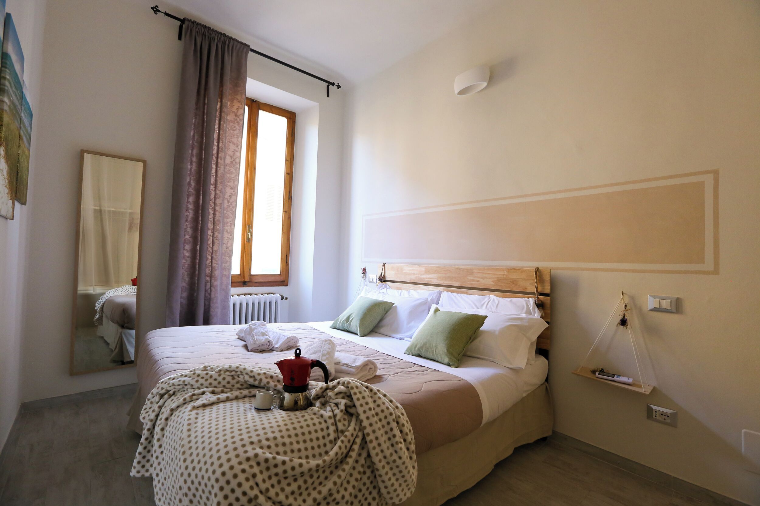 Una cama bien hecha con cabecera de madera, una mesita de noche con lámpara, una ventana con cortinas y un estante en la pared con un control remoto.