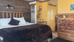 A bedroom with a wooden headboard, a bed with white and dark brown bedding, a small bedside table with a lamp, and a door leading to another room.