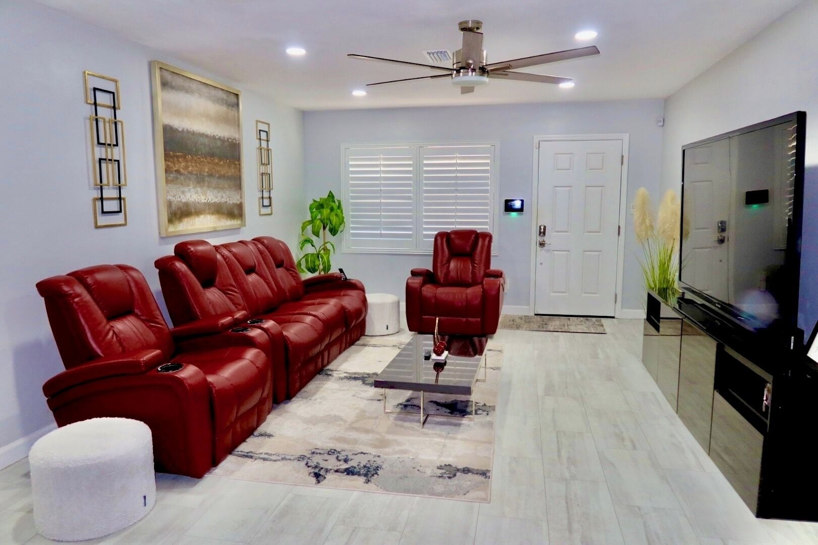 Un salon avec un canapé en cuir rouge, un fauteuil inclinable rouge, un ventilateur de plafond, un téléviseur à écran plat et une grande fenêtre avec des stores.