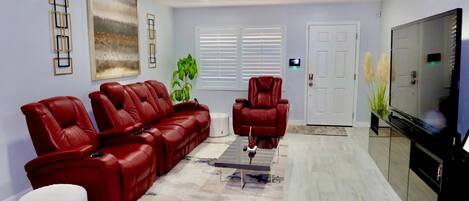 A living room with a red leather sofa, a red recliner, a ceiling fan, a flat-screen TV, and a large window with blinds.