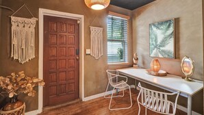 A room with a wooden door, a white desk with two chairs, a hanging macrame wall decoration, a framed picture, and a pendant light. - Chic Boho Treehouse Studio in Morningside (Miami)