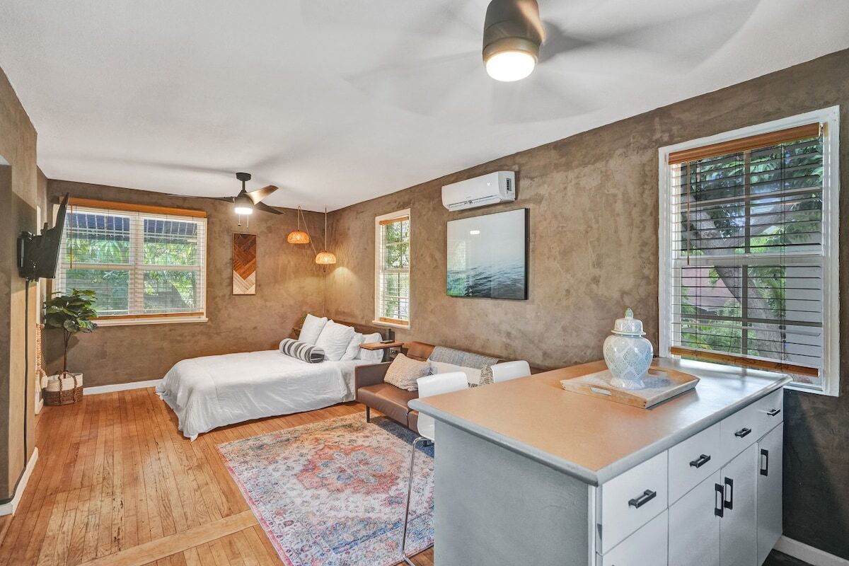 Une chambre à coucher comprenant un lit, un canapé, un bureau avec des tiroirs, un ventilateur de plafond et une grande fenêtre avec des stores.