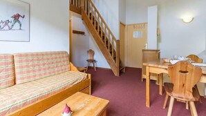Una habitación acogedora con una escalera de madera, un sofá, una mesa de comedor con sillas y un pequeño taburete.