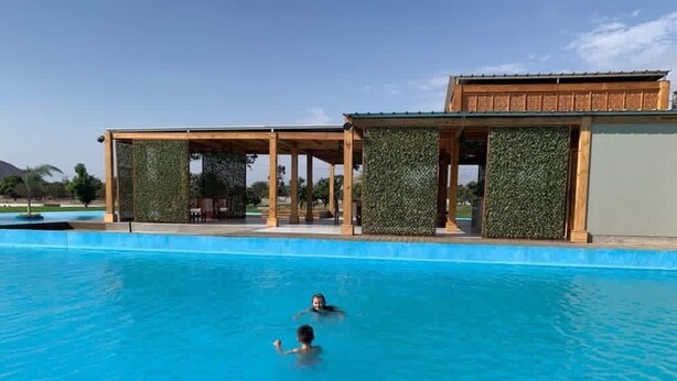 A swimming pool with two people swimming, surrounded by wooden structures with green walls. - Sol y Relax Family Home in Huacachina, Ica (Ica)