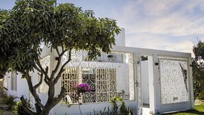A white building with a decorative metal gate, a tree in front, and a clear sky. - Sol y Relax Family Home in Huacachina, Ica (Ica)