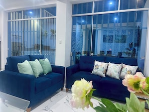 A modern living room with two blue sofas, a glass partition, and a view of the outside.