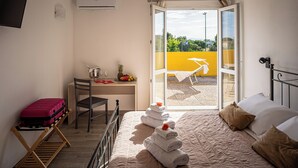 Habitación de hotel con cama, escritorio, silla, maleta y balcón con pared amarilla y una silla.