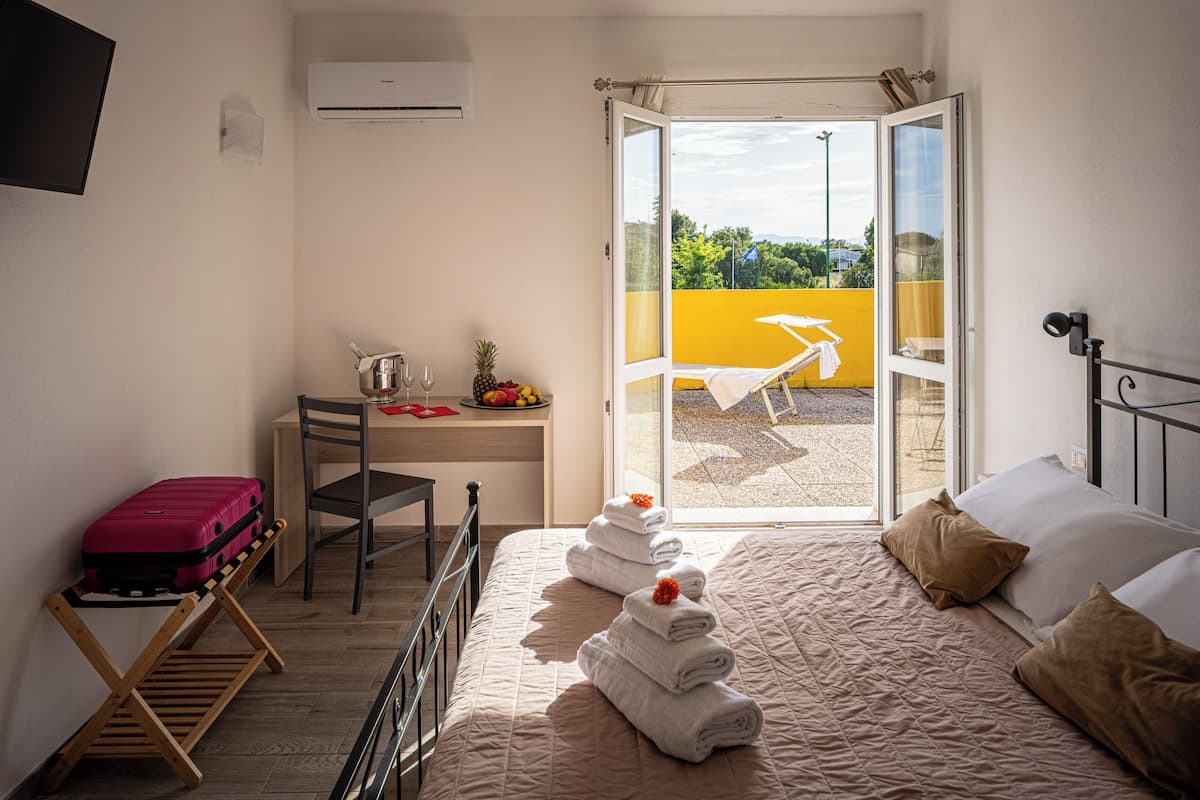Habitación de hotel con cama, escritorio, silla, maleta y balcón con pared amarilla y una silla.