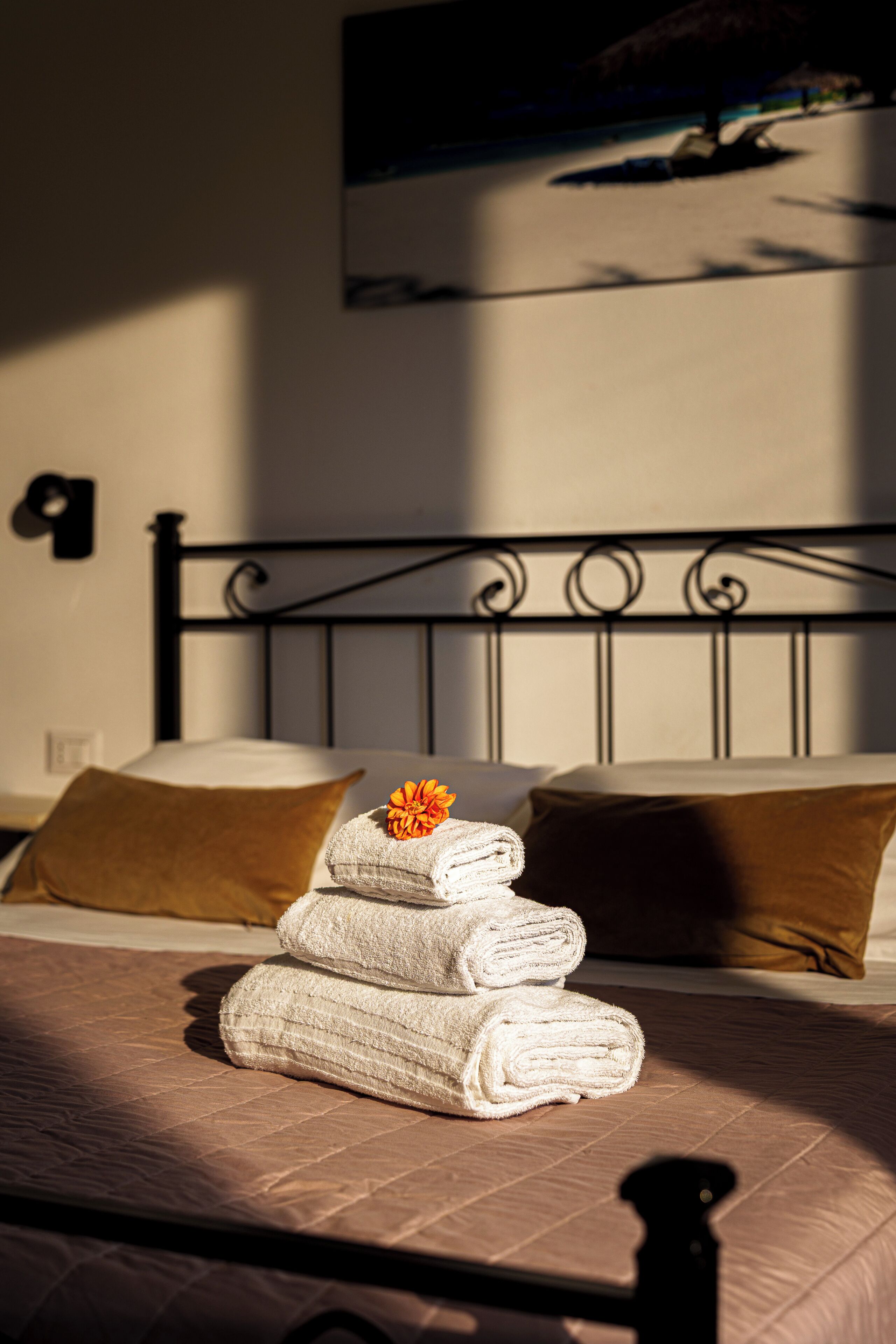 A bed with rolled white towels and a single orange flower, a brown pillow, and a metal headboard.
