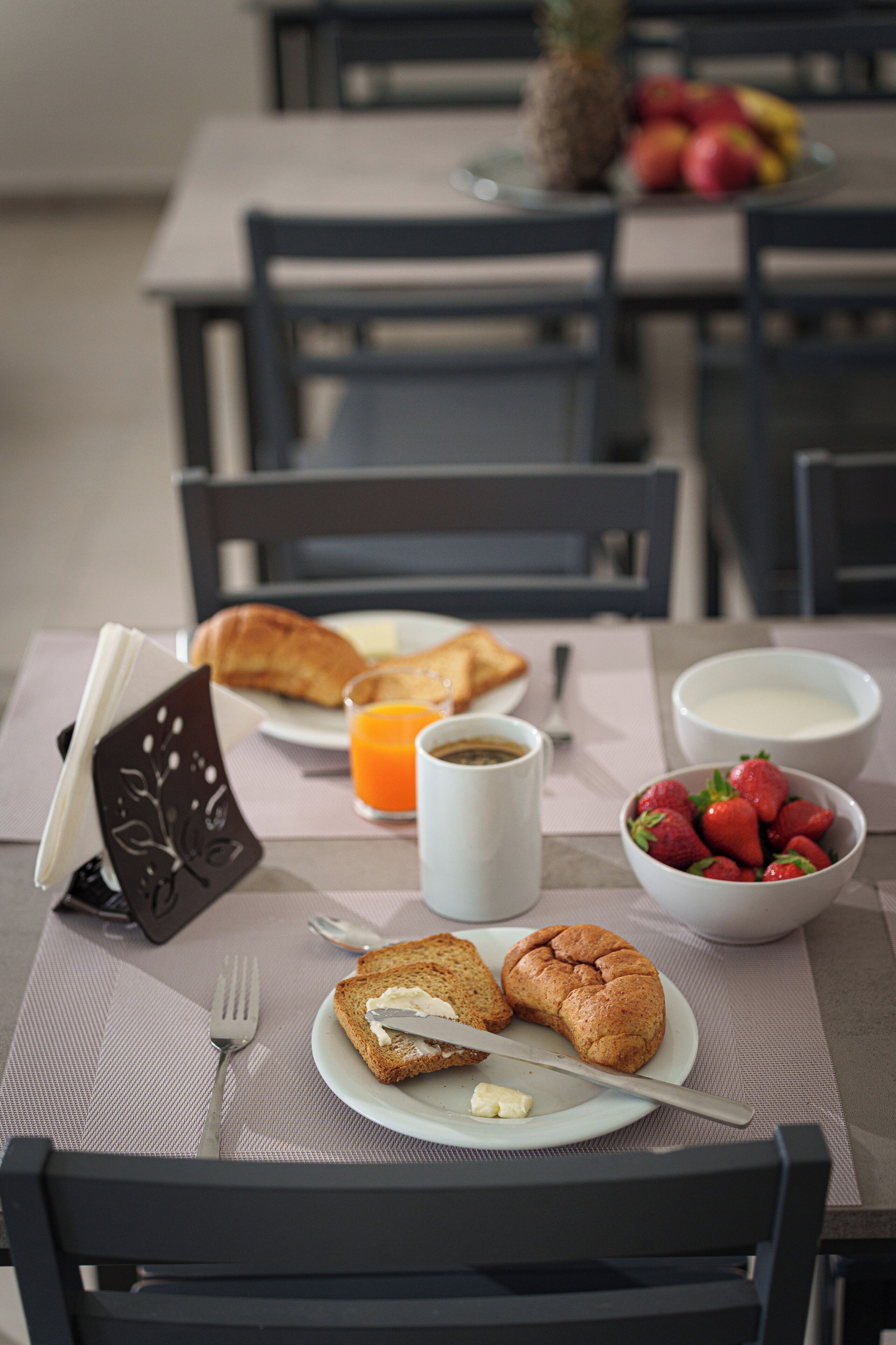 Et morgenbord med ristet brød, croissanter, en skål jordbær, et glas appelsinjuice og en kop kaffe på et bord.
