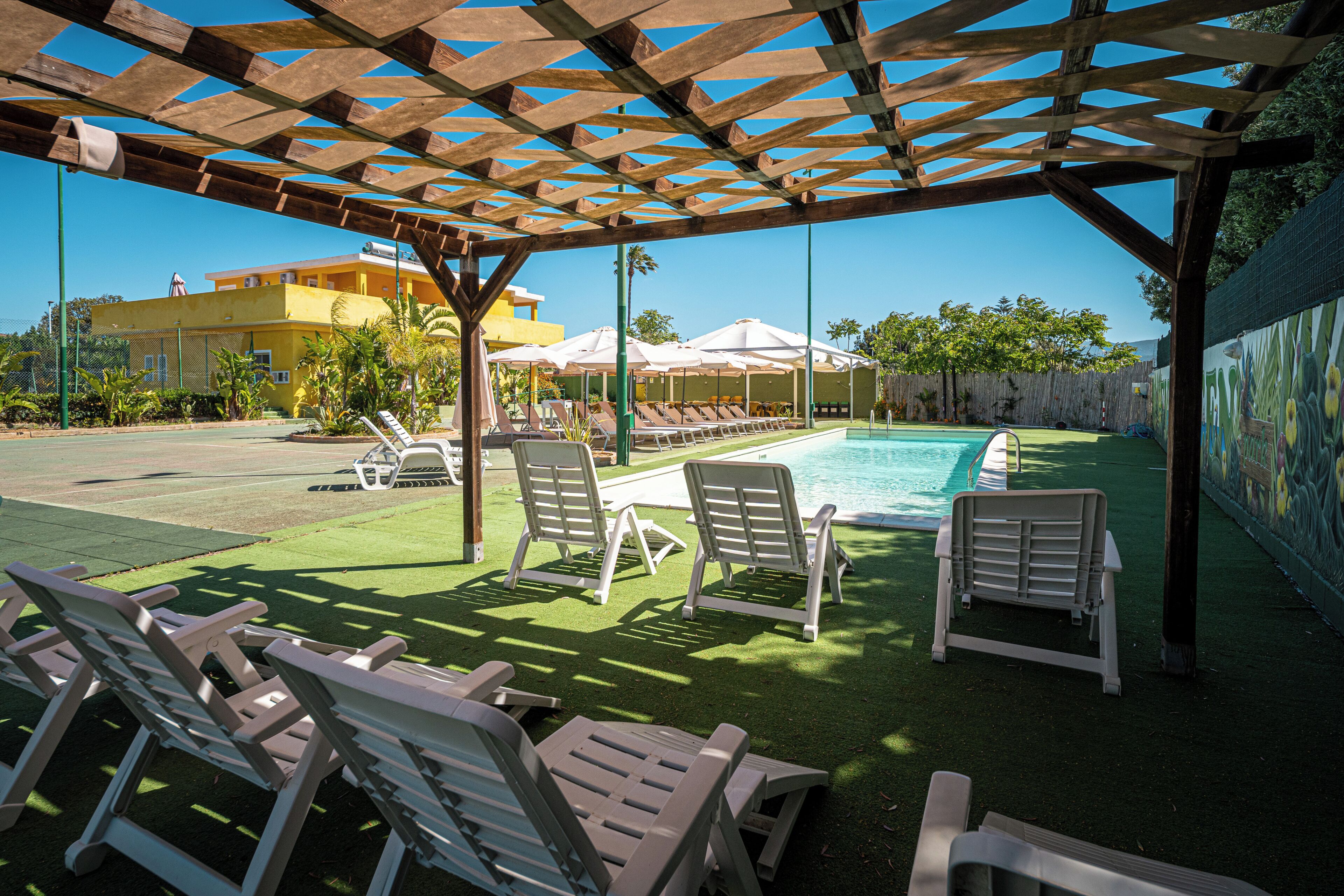 Ein Poolbereich mit Liegestühlen und Sonnenschirmen, eine Holzpergola und ein Gebäude im Hintergrund.