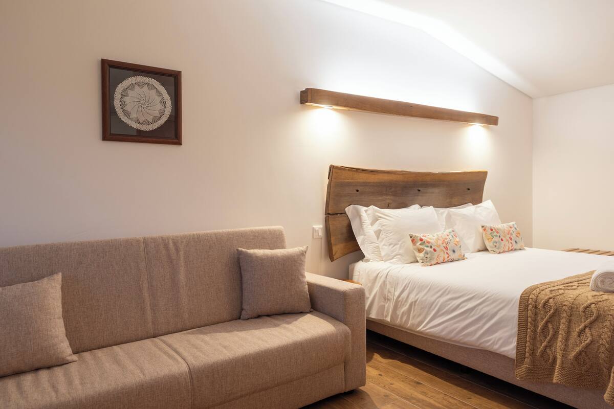 A bedroom with a wooden headboard, a beige sofa, a white bed with colorful pillows, and a framed artwork on the wall.