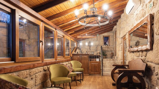 A cozy room with wooden beams, stone walls, and a chandelier. There are two green chairs, a wooden table, and a stone fireplace. - Quinta do Limite Agroturismo (Covilhã)