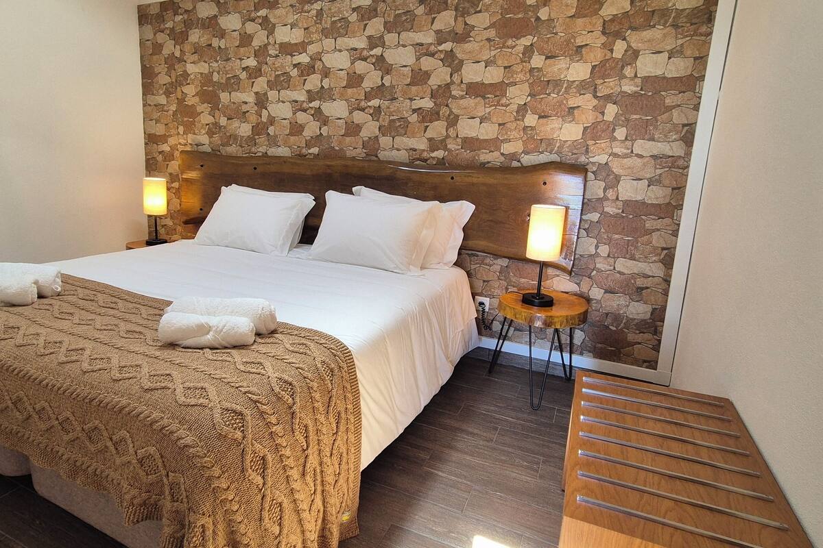 A neatly made bed with a patterned blanket and two towels, a wooden headboard, stone wall, and a small round table with a lamp.