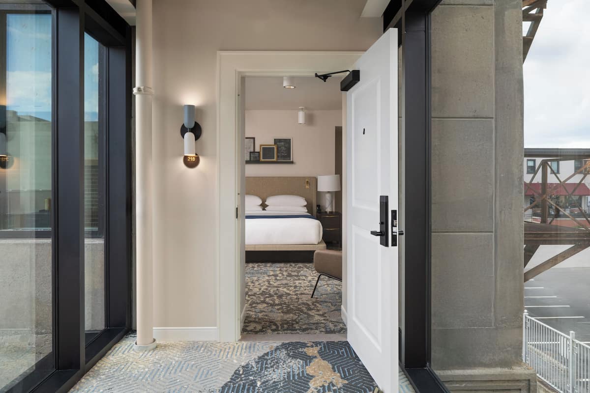 Habitación de hotel con cama, mesita de noche, lámpara y vistas al exterior.