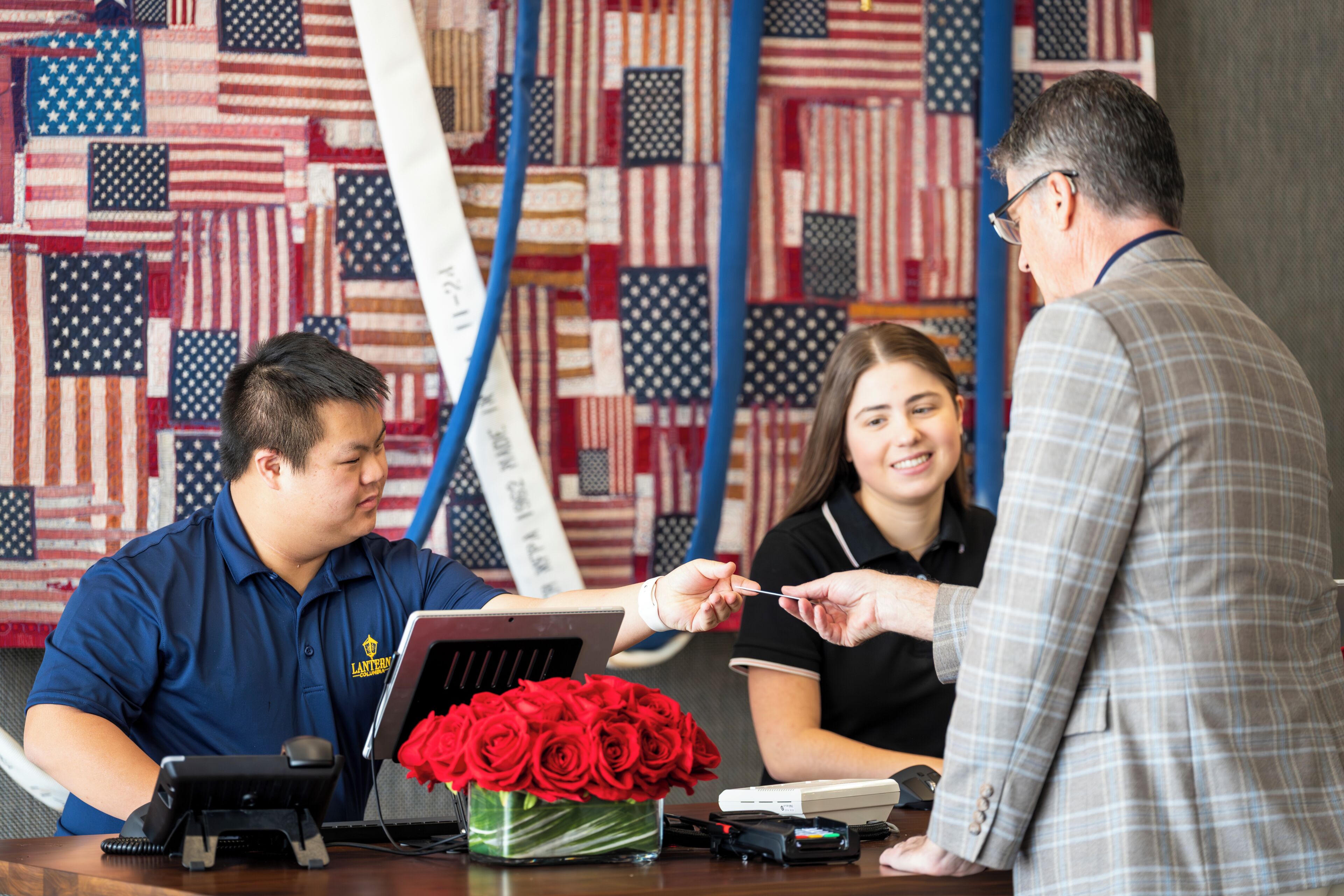 A reception desk with a laptop, a vase of red roses, and a flag wall in the background.