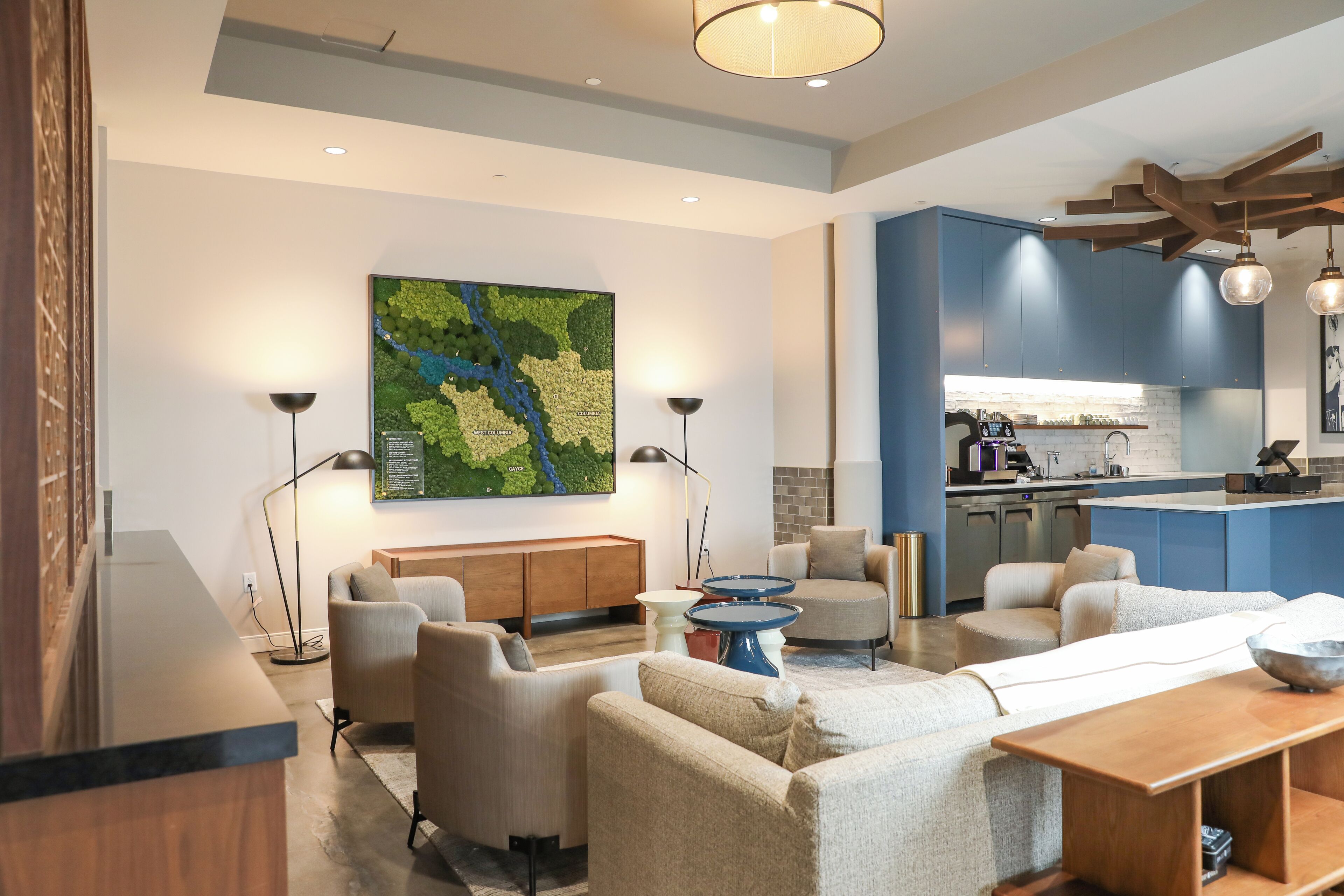A modern living room with a sofa set, a coffee table, and a dining table. There is a kitchen area with blue cabinets and a hanging light fixture. A map is displayed on the wall.