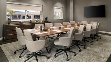 A conference room with a long table, multiple chairs, a TV on the wall, and a window with blinds.