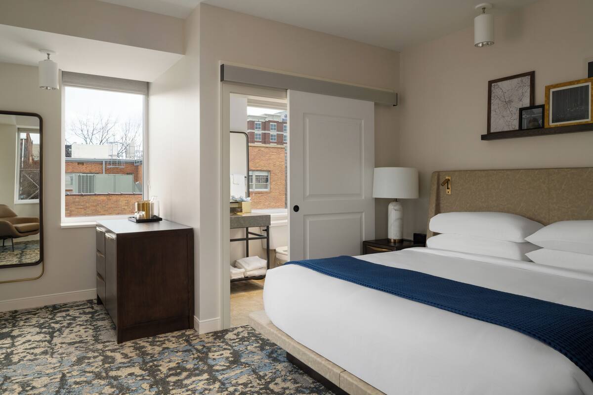 Habitación de hotel con cama, mesita de noche, lámpara y vista a un edificio de ladrillo a través de una puerta abierta.