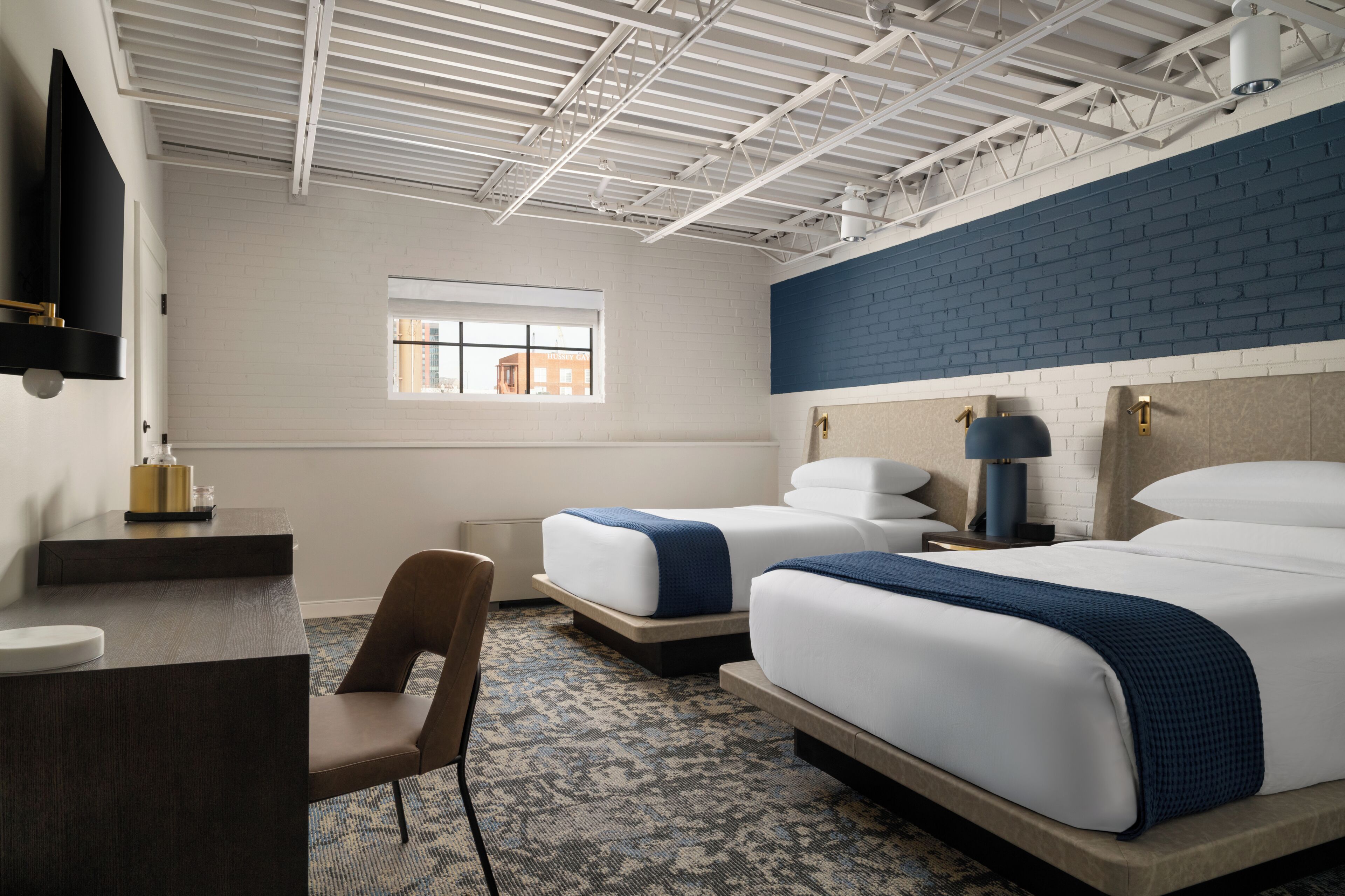 A hotel room with two beds, a desk, a chair, a window, and a brick accent wall.