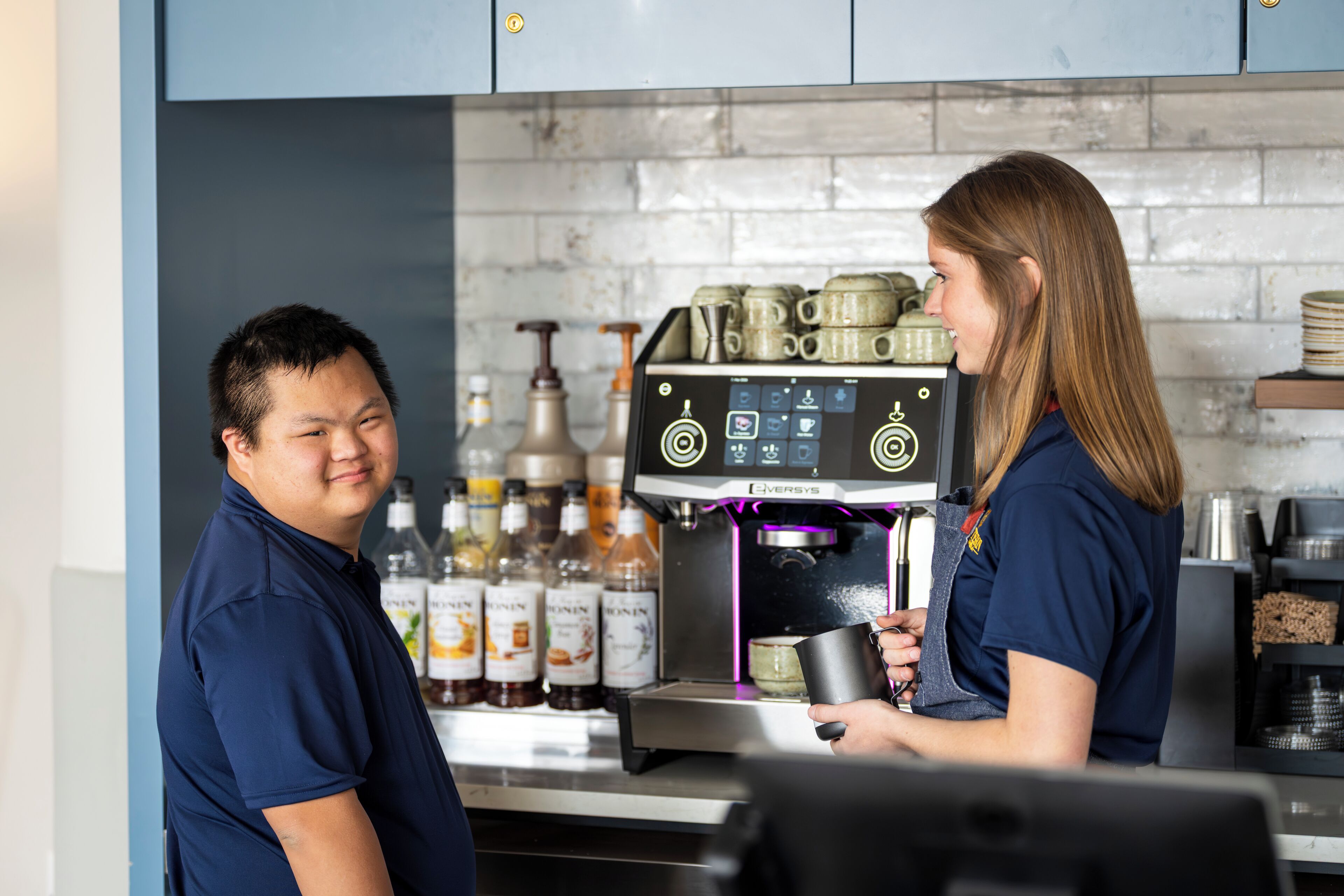 A barista preparing a drink at a coffee machine in a kitchen setting with shelves of bottles.