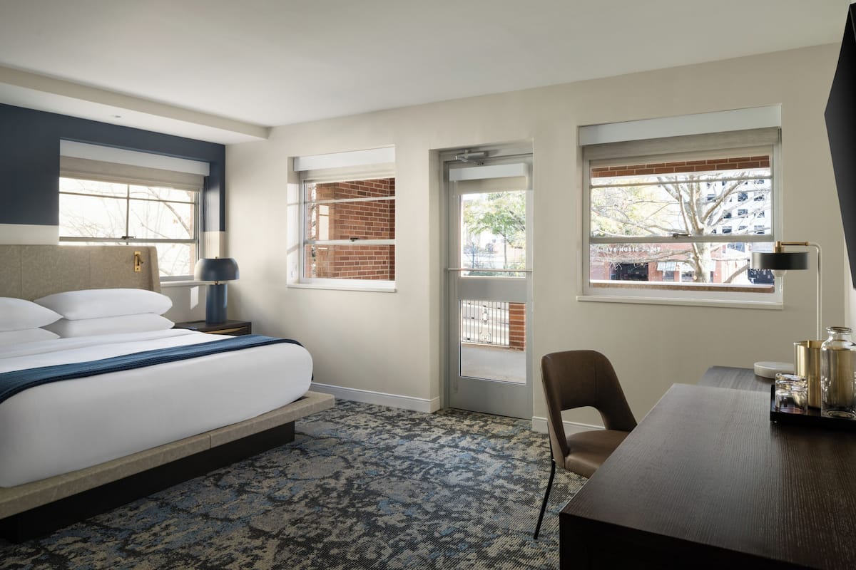 Habitación de hotel con cama, escritorio, dos ventanas, una puerta y alfombra estampada.