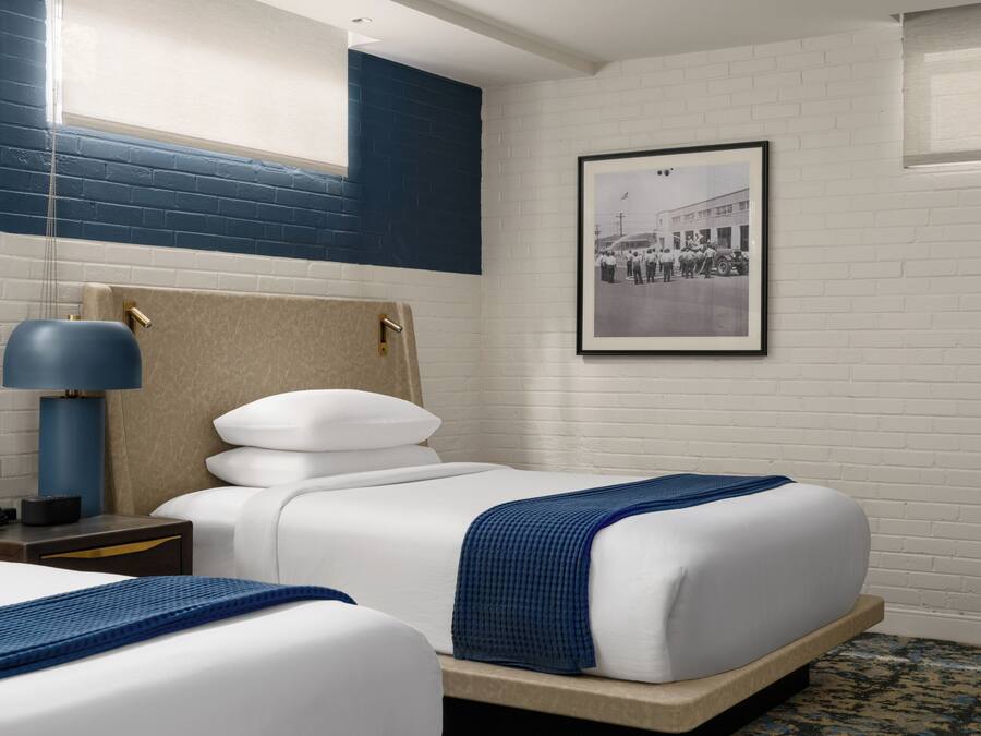 A hotel room with a bed, a nightstand with a lamp, a framed picture on the wall, and a window with blinds.