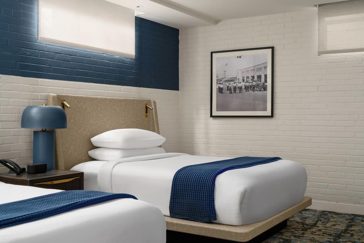 Una habitación de hotel con dos camas, una lámpara azul, un cuadro en la pared y una ventana con vista a un edificio.
