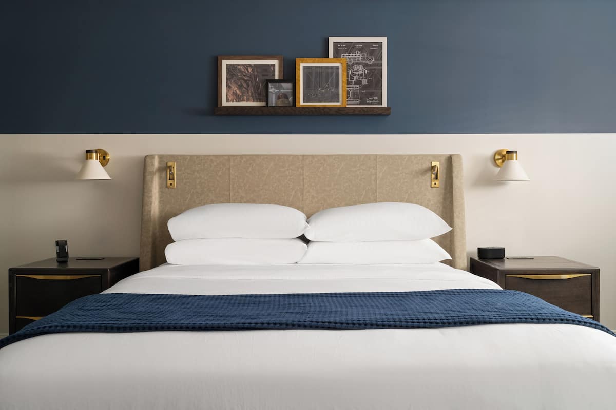 Una cama bien hecha con sábanas blancas, un plaid azul y un cabecero con detalles dorados. Hay dos mesitas de noche, cada una con una lámpara y un pequeño objeto encima. Encima de la cama, hay tres cuadros enmarcados sobre una estantería.