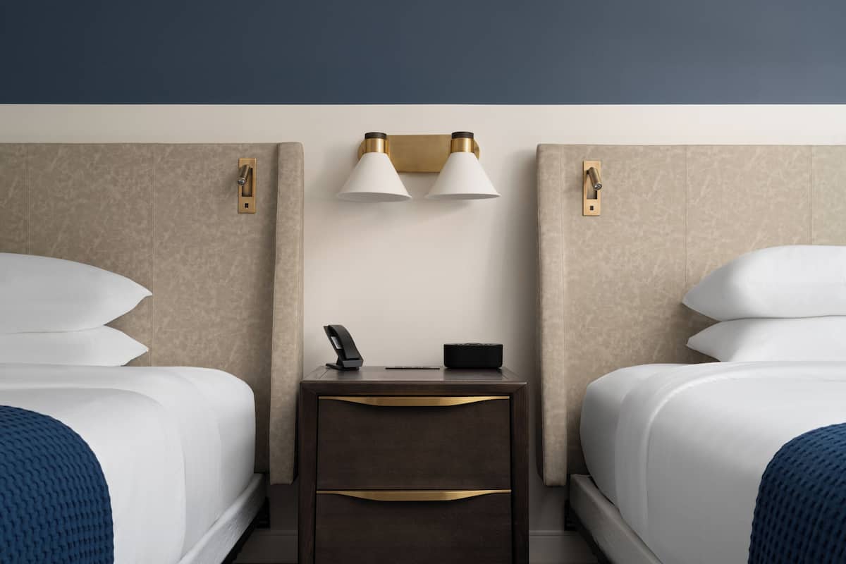 Habitación de hotel con dos camas, una mesita de noche con lámpara, teléfono y una pequeña mesa.