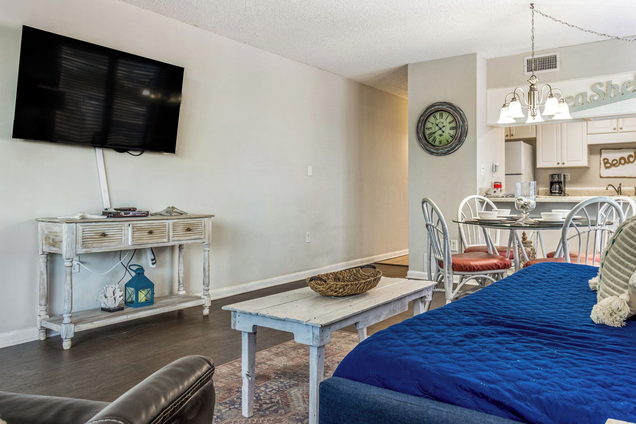 Un salon avec un téléviseur, une table à manger avec des chaises, un canapé bleu et un coin cuisine avec une horloge.