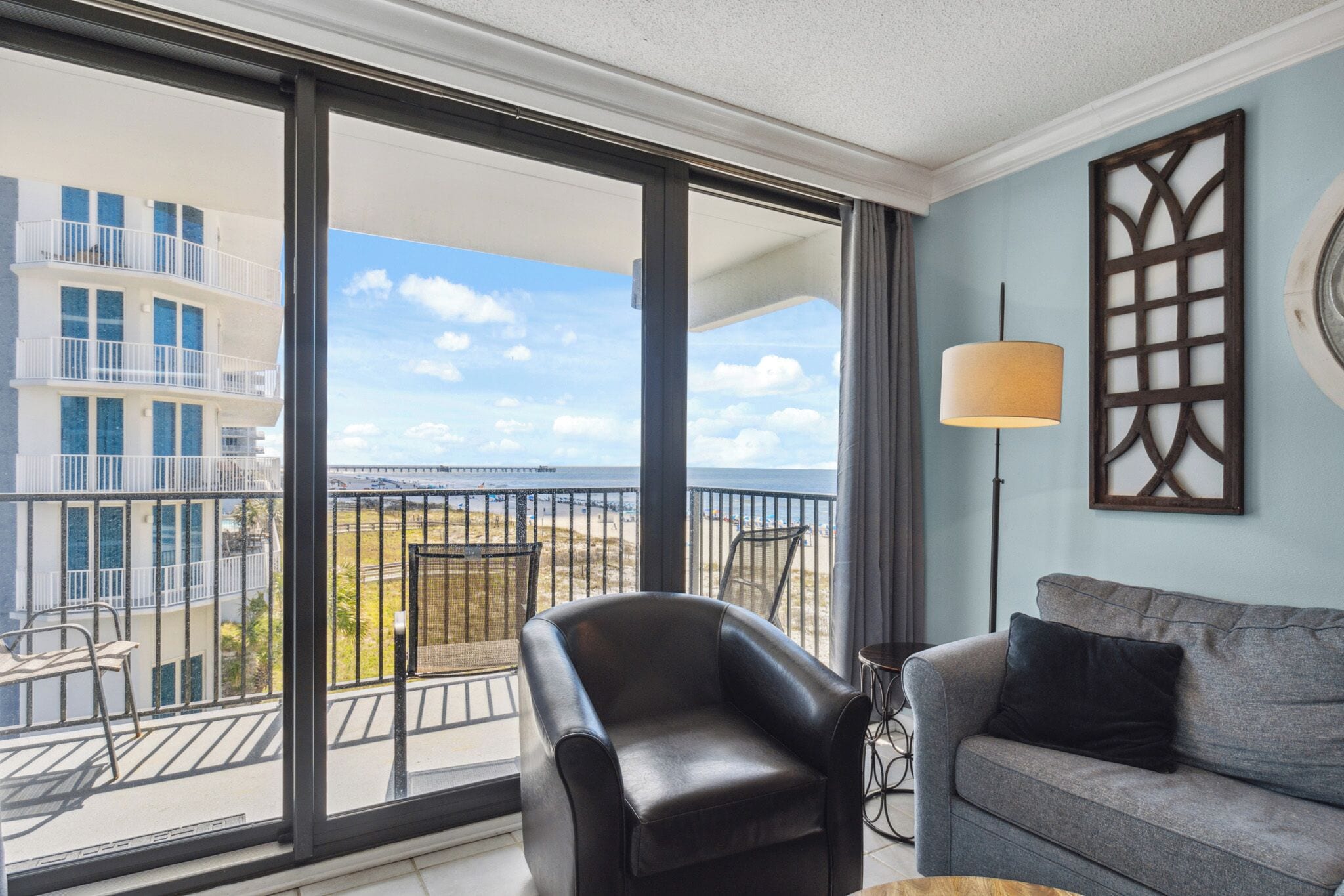 A balcony with a view of a building and beach, a black leather chair, a grey sofa, and a floor lamp.