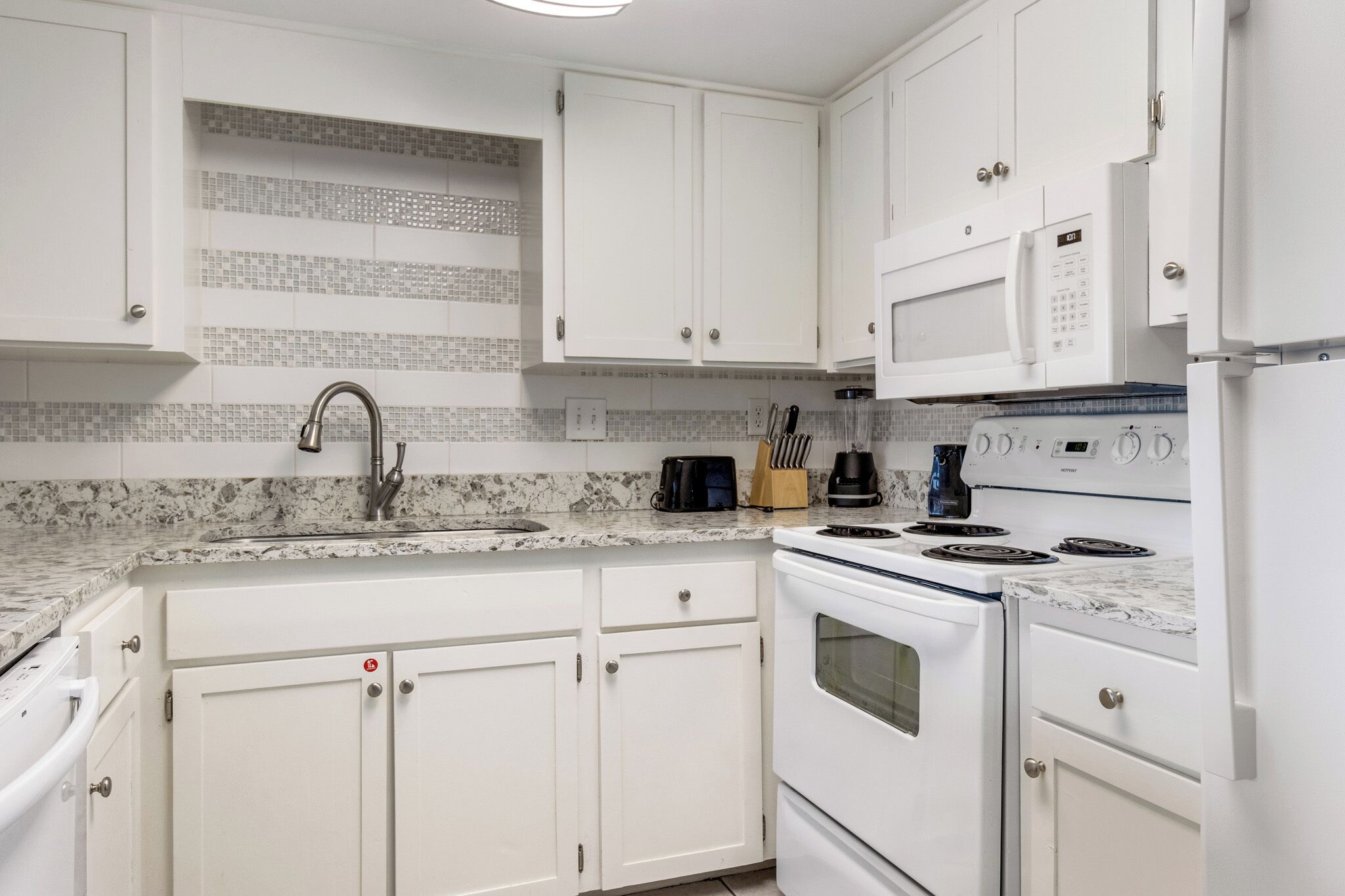 A kitchen with white cabinets, a granite countertop, a stainless steel sink, a built-in microwave, a stove, and a coffee maker.