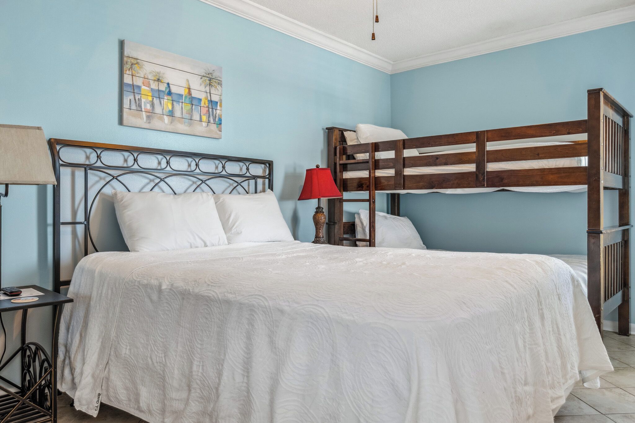 A room with a bunk bed, a single bed with white bedding, a red lamp, and a painting on the wall.