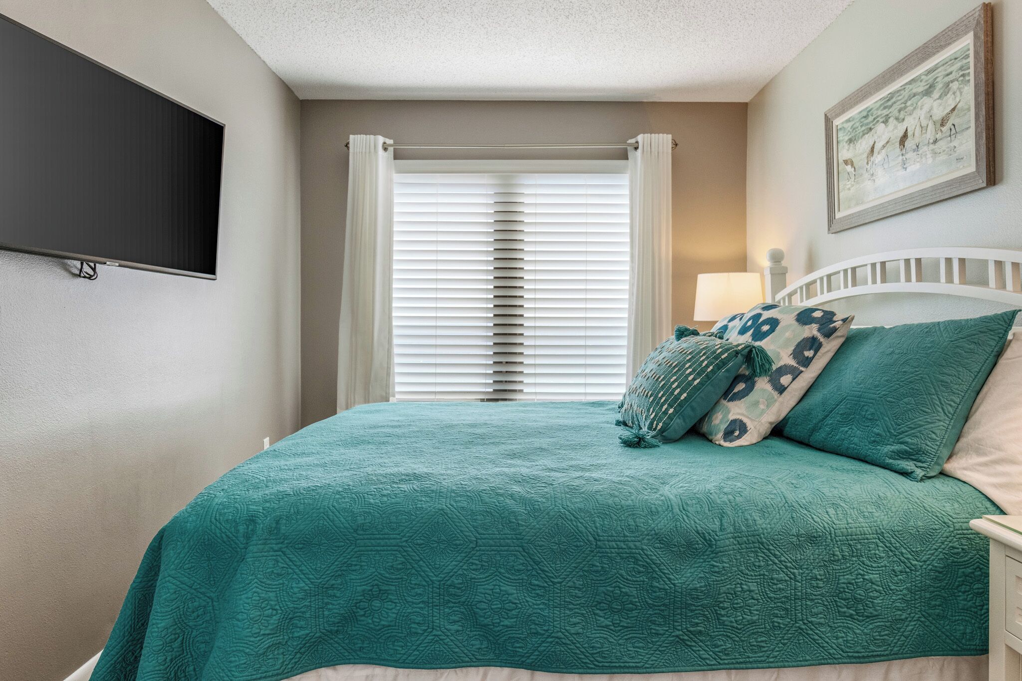 A bedroom with a bed, a TV mounted on the wall, a window with blinds, and a painting on the wall.