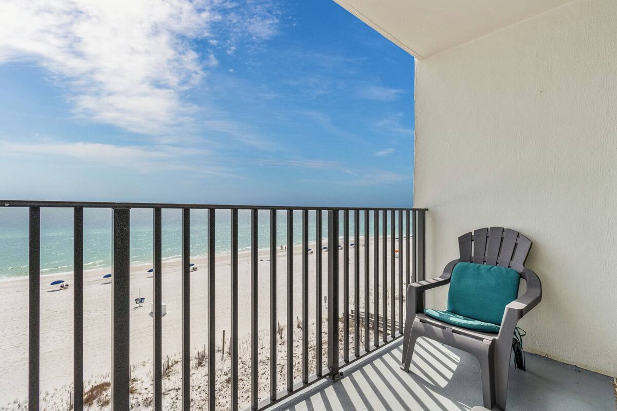 A balcony with a view of a beach, a grey Adirondack chair, and a white wall.