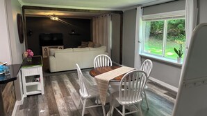 A dining area with a table and chairs, a sofa in the background, and a window with a view of greenery.