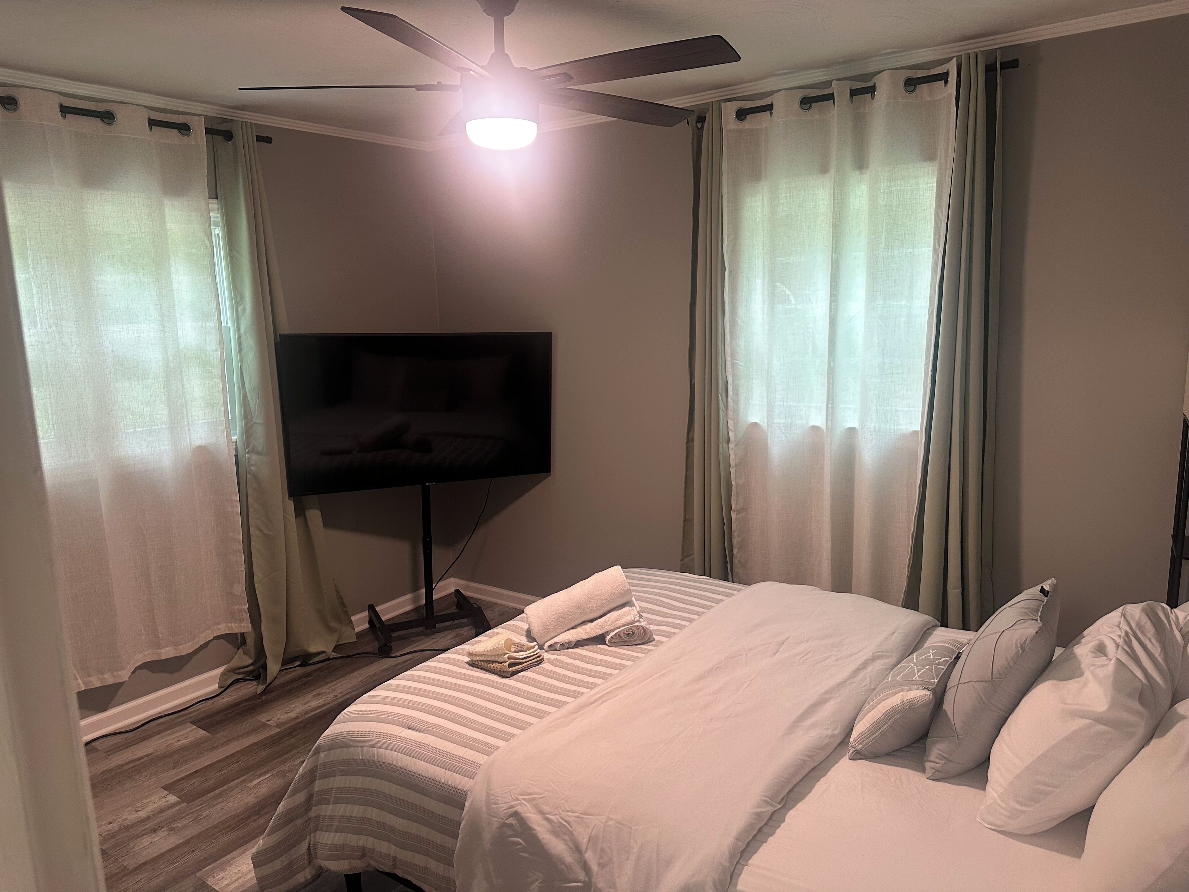A hotel room with a bed, a flat-screen TV mounted on the wall, a ceiling fan, and sheer curtains.